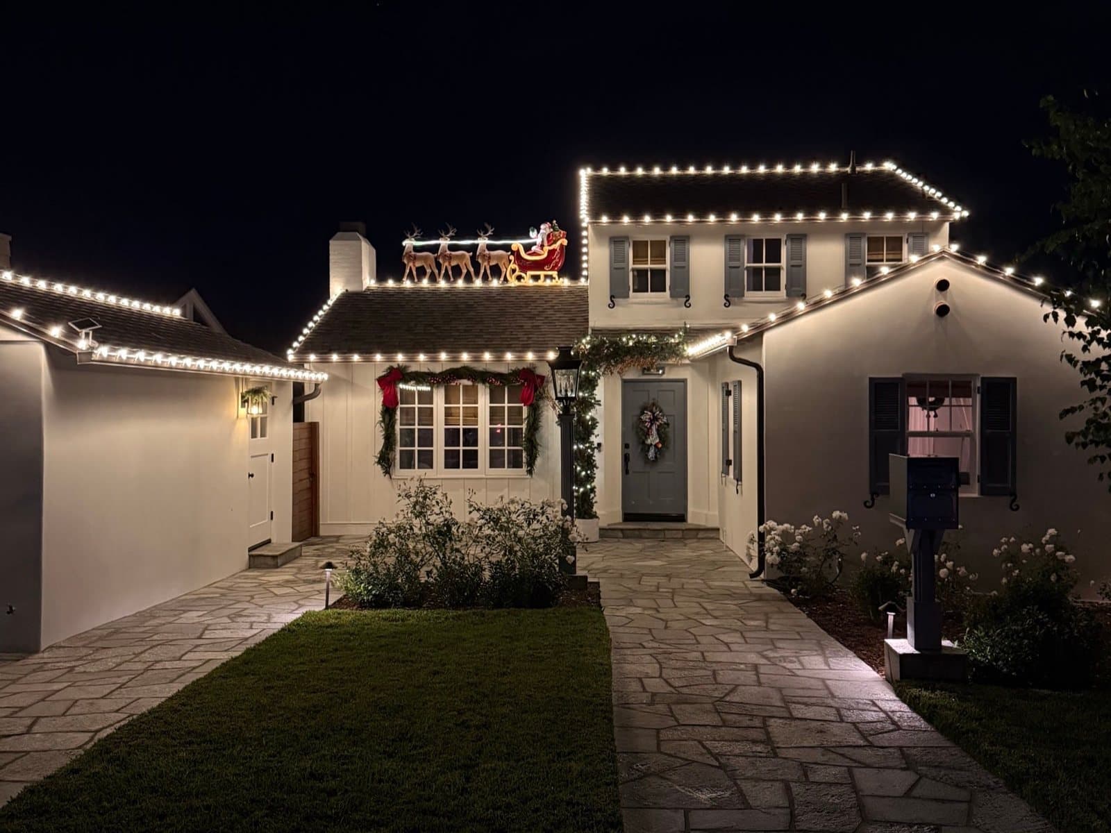 The same La Jolla cottage on install night: warm white C9 bulbs lit along every roofline, garland and bows on the front window, a wreath on the door, and a lit Santa sleigh with reindeer on the roof